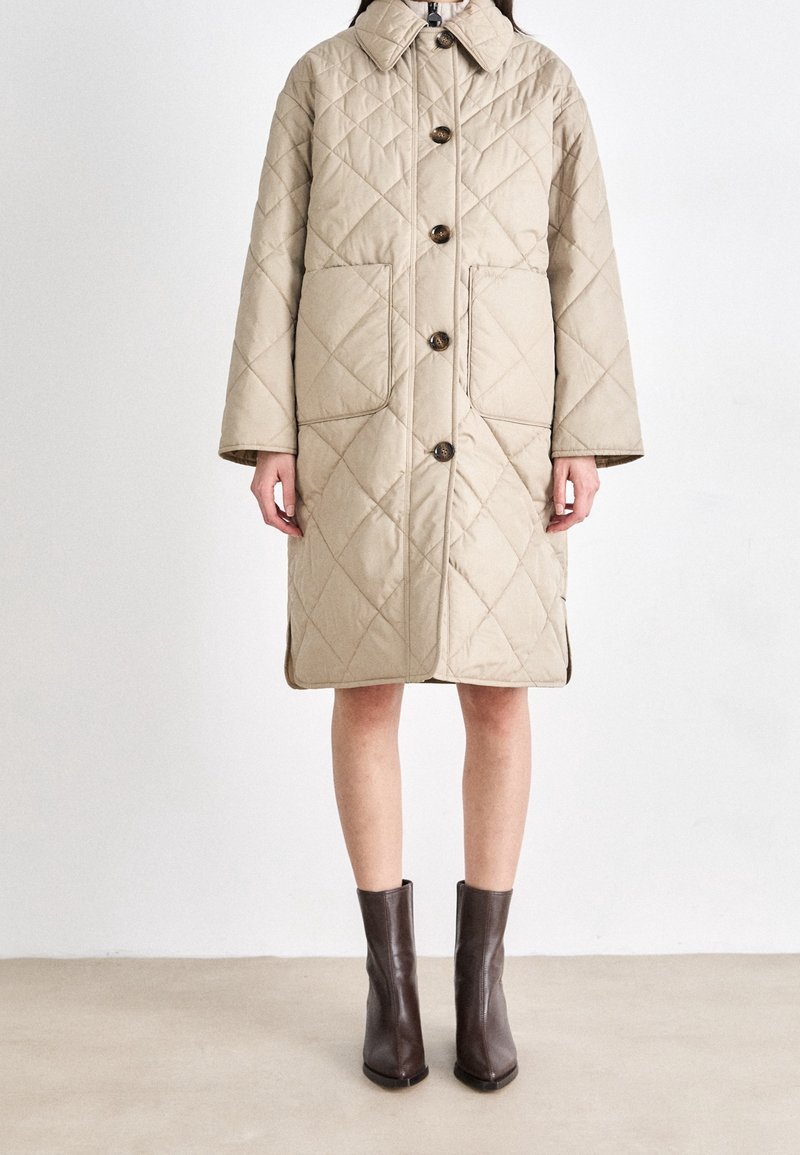 Manteau beige matelassé jusqu'aux genoux avec boutons sur le devant et grandes poches, porté avec des bottines marron foncé sur un fond neutre.