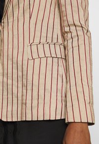 Blazer beige con strisce verticali rosse, tessuto testurizzato, colletto a punta e una tasca sul petto con dettaglio a patta.