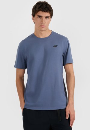Młody mężczyzna w prostym niebieskim t-shircie z krótkim rękawem z małym czarnym logo "4F" na lewej piersi, stojący na jednolitym tle.