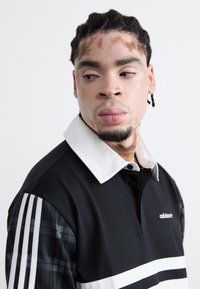 Black polo shirt with white collar and horizontal stripe. Features plaid patterned sleeves and an embroidered logo on the chest.