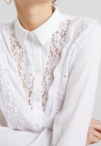 Weißes Hemd mit Knopfleiste und einem Spitzenbesatz auf der Vorderseite. Der Stoff hat eine glatte Textur, und der Kragen ist strukturiert mit langen Ärmeln.
