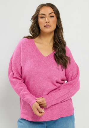 Femme aux longs cheveux bruns portant un pull rose vif à col en V et un jean bleu, debout devant un fond clair uni.