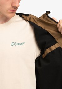 Chaqueta negra con acentos marrones, parcialmente abierta para revelar una camiseta crema con texto en color teal que dice "Element". Material de algodón, diseño casual.