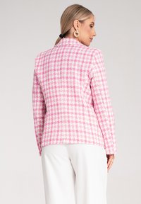 Różowo-biała marynarka w wzór houndstooth o dopasowanym kroju, teksturowanej tkaninie i wykrojonym kołnierzu; zestawiona z białymi spodniami.