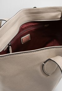 Sac cabas synthétique beige avec une finition texturée, doté d'un intérieur rouge profond et d'une poche intérieure zippée. Étiquette logo enduite visible.