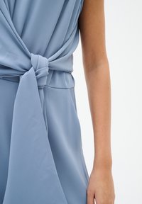 Tuta azzurro chiaro con un design avvolto in vita, caratterizzata da un dettaglio a nodo e una texture di tessuto morbido. Sono visibili maniche corte.