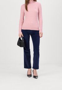 Pull à col roulé en maille torsadée rose, pantalon en velours côtelé bleu marine, sac à main noir et escarpins noirs à bouts arrondis. Design simple et ajusté.
