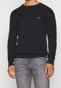 Maglione nero a maglia con scollatura a girocollo, polsini e bordo a coste, presenta un piccolo etichetta con logo sul davanti. Abbinato a jeans grigi.