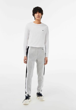 Chemise blanche à manches longues, pantalon de survêtement gris avec des panneaux latéraux noirs et des rayures blanches, associé à des baskets blanches. Design simple et athlétique.