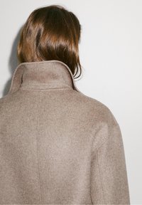 Beige uldfrakke med høj krave, synlig tekstur og enkelt design. Bagsiden har rene linjer og sømme.