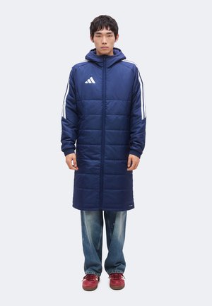 Giovane uomo che indossa un lungo cappotto trapuntato blu navy con strisce bianche sulle maniche, jeans blu e scarpe da ginnastica rosse, in piedi davanti a uno sfondo semplice.