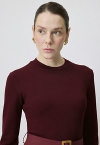 Burgundy långärmad topp med rund halsringning, kombinerad med ett brett bordeaux bälte med en guldig rektangulär spänne och guldaccentörhängen.