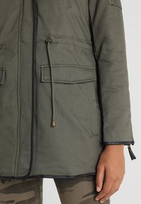 Veste vert olive avec fermeture éclair à l'avant, poche à rabat avec cordon de serrage, portée avec un pantalon camouflage, montrant le bras et la main droits.