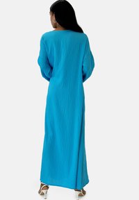 Lang, blå maxikjole i tekstureret stof med lange ærmer og en afslappet pasform. Har en let slids på den ene side og en rund halsudskæring.