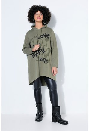 Kvinde med krøllet hår iført en olivengrøn oversized hættetrøje med sort tekst, sorte læderleggings og sorte ankelstøvler, stående.