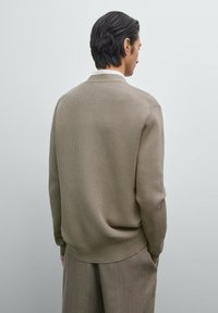 Beige gestrickter Pullover mit einem strukturierten Muster. Das Design verfügt über einen runden Ausschnitt und eine gerippte Manschette, kombiniert mit grauen Hosen.