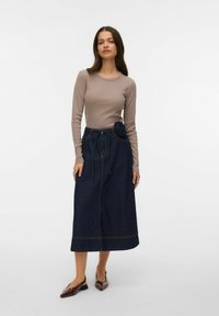 Beige top met lange mouwen gecombineerd met een donkere denimrok met een bloemdetail bij de taille, zichtbaar stiksel en een vloeiende vorm.