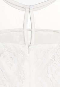 Vit klänning med spetsöverlägg med blommönster, meshaxelparti, knappstängning i ryggen och zippralj detalj vid midjan.