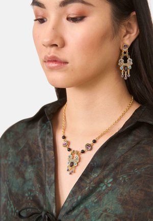 Mujer con un collar de oro ornamentado y pendientes a juego con gemas púrpuras y azules, vestida con una blusa oscura de estampado.