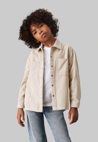 Camicia button-up in corduroy crema con due tasche sul petto, indossata sopra una t-shirt bianca, abbinata a jeans azzurri chiari; tessuto testurizzato visibile.