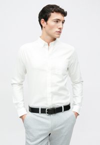 Weißes Hemd mit langen Ärmeln und Brusttasche, kombiniert mit hellgrauen karierten Hosen und einem schwarzen Gürtel.