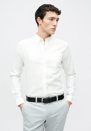 Λευκό πουκάμισο με κουμπιά, μακριά μανίκια και τσέπη στο στήθος, συνδυασμένο με ανοιχτό γκρι καρό παντελόνι και μαύρη ζώνη.