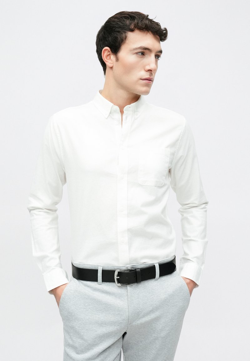 Λευκό πουκάμισο με κουμπιά, μακριά μανίκια και τσέπη στο στήθος, συνδυασμένο με ανοιχτό γκρι καρό παντελόνι και μαύρη ζώνη.