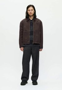 Jovem de pé, virado para a frente, usando uma jaqueta de fecho éclair castanho escuro, uma camisa listrada em preto e cinza, calças largas escuras e sapatos pretos, contra um fundo branco.