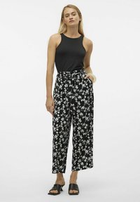 Zwarte mouwloze top gecombineerd met hoge, wijde broek met een wit bloemenpatroon. Model draagt zwarte instapschoenen.