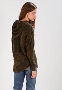 Oliven grønn, oversized hettegenser med myk stoff, lange ermer og snor i hetta; har et subtilt kamuflasjemønster.