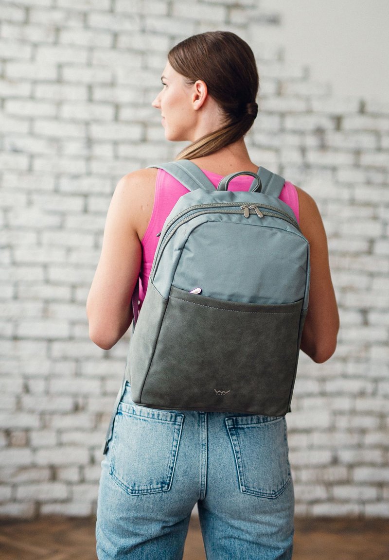 Grauer Stoff-Rucksack mit einem dunkler grauen Boden, zwei gepolsterten Schultergurten, einem oberen Griff und Reißverschlusstaschen. Muster und Texturen sind sichtbar.
