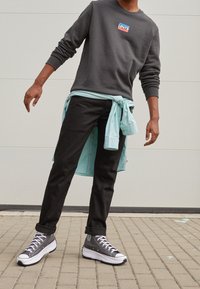 Osoba v tmavě šedé mikině Levi's, černých džínech, šedých vysokých teniskách a s světle modrou košilí uvázanou v pase stojí na chodníku.