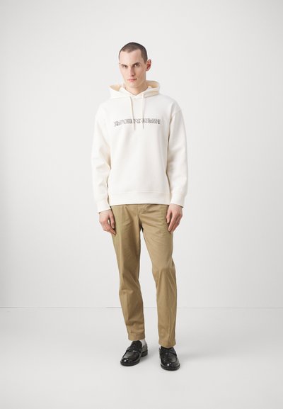 Felpa bianca con cappuccio e logo frontale, abbinata a pantaloni marrone chiaro e scarpe nere. Il modello è appoggiato a uno sfondo semplice.
