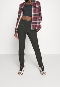 Pantalones negros ajustados con una textura suave, bolsillo con cremallera funcional y una cintura de altura media. Combinados con una camisa de cuadros y zapatillas deportivas.