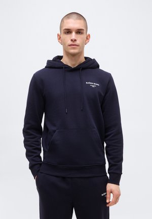 Giovane uomo con capelli corti che indossa una felpa con cappuccio blu navy Björn Borg e pantaloni abbinati, in piedi contro uno sfondo chiaro e semplice.