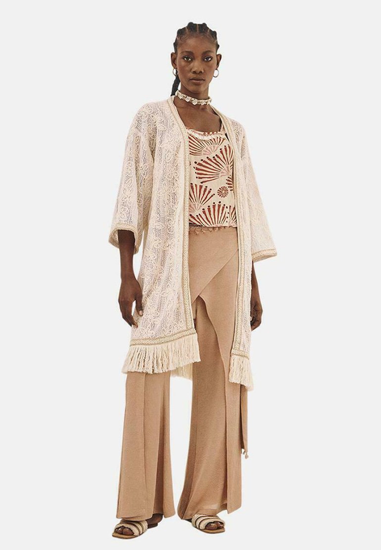 Kimono de encaje beige con flecos, superpuesto sobre una blusa estampada y pantalones palazzo beige. Accesorios con un choker y sandalias.