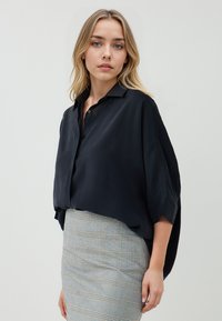 Blouse noire à col, décontractée et à manches courtes, associée à une jupe ajustée en tartan gris. Texture lisse, détails minimalistes.