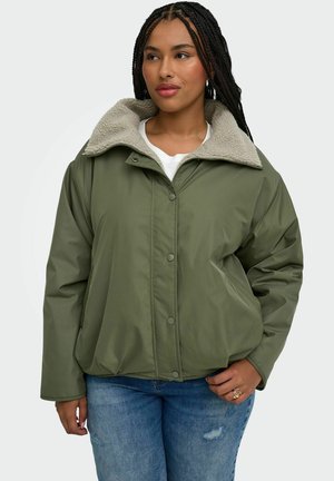 Femme aux cheveux tressés portant une veste vert olive avec un col en polaire beige et un jean bleu, debout avec une main dans la poche.