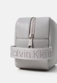 Pochette zippée en cuir gris avec une surface texturée, des bords arrondis et une sangle en tissu embossée du logo. Détail en métal argenté pour la fermeture éclair.