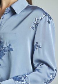 Blusa in satin blu chiaro con decorazioni floreali in paillettes di tonalità più scure. Include un colletto classico e chiusura a bottoni.