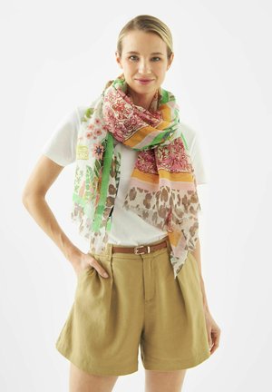 Frau trägt einen bunt gemusterten Schal, weißes T-Shirt, beige hochtaillierte Shorts mit Gürtel, steht mit einer Hand in der Tasche.