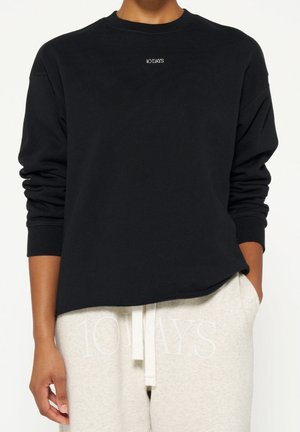 Person trägt ein schwarzes Langarmshirt und eine hellbeige Jogginghose, beide mit dem Aufdruck "10DAYS", die Hände locker an den Seiten.