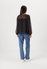 Blouse noire transparente à motif de points argentés, manches longues, avec un détail dos en forme de trou de serrure, associée à un jean bleu clair à coupe ample et des baskets blanches.