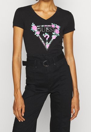 Kobieta w czarnej koszulce z dekoltem w serek z kwiatowym logo Guess oraz czarnych spodniach z wysokim stanem, z materiałowym paskiem z srebrną klamrą w kształcie litery D.