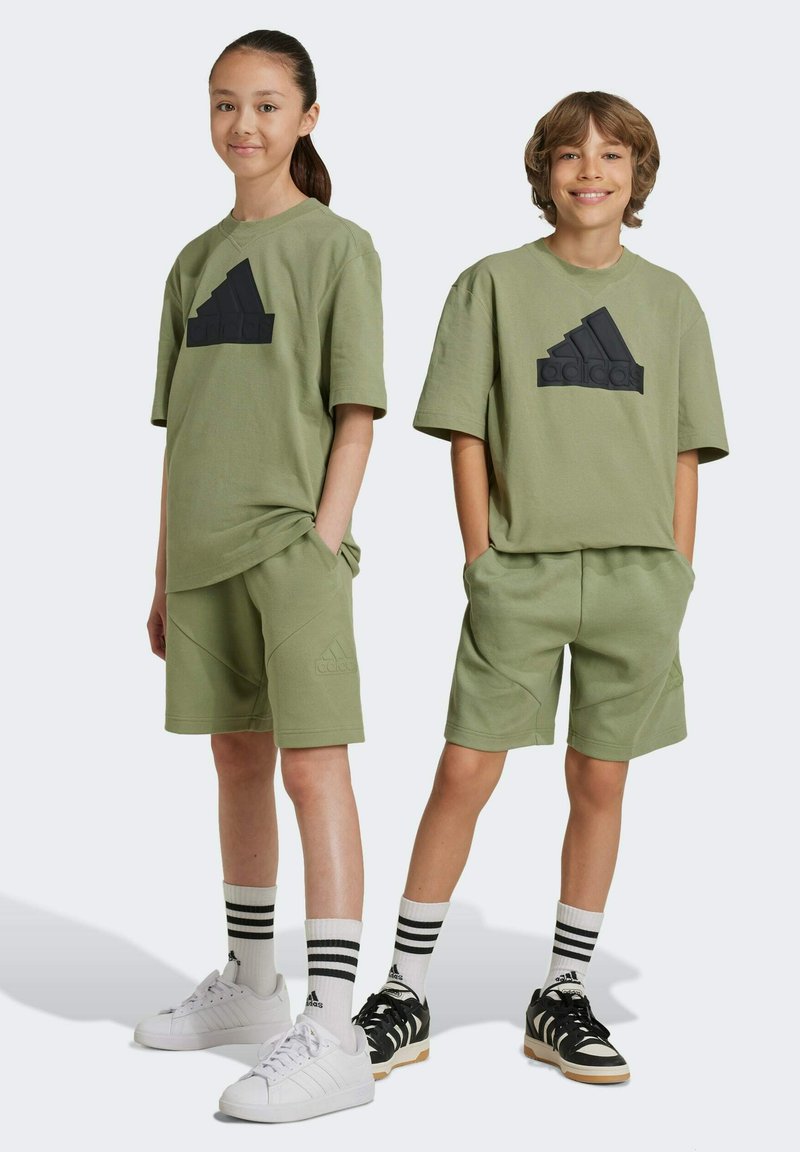 T-shirt e pantaloni corti di cotone verde oliva, con il logo Adidas nero. Sneakers bianche con dettagli neri, calze a righe. Design semplice.