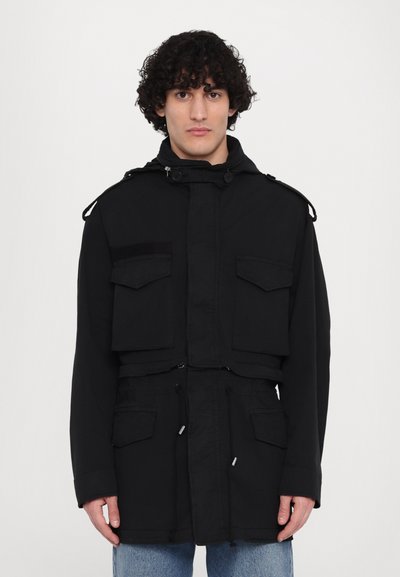 Veste militaire noire avec un col montant, poches avant, taille à cordon de serrage et détails en boutons sur les épaules. Le tissu semble robuste et texturé.