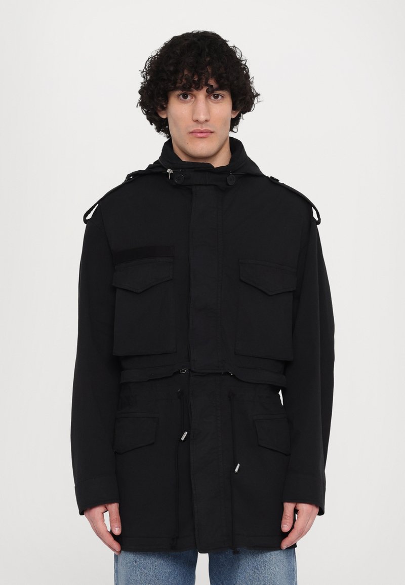 Zwarte militaire stijljack met een hoge kraag, voorzakken, aansnoerbare taille en knopendetails op de schouders. De stof lijkt stevig en gestructureerd.