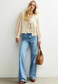Blonde Frau trägt eine blumige Bluse, eine beigefarbene Knopfweste, weite blaue Jeans, braune Sandalen und hält eine hellbraune Lederhandtasche, während sie gegen eine weiße Wand steht.