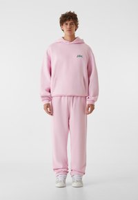 Rozā kapuce ar priekšējo kabatu un izšūtu logotipu, apvienota ar atbilstošām rozā sporta biksēm. Mīksts audums un brīvas formas dizains.