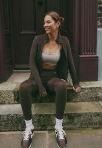 Young woman in brown activewear and sneakers sitting on stone steps, smiling and looking to the side by a dark wooden door.